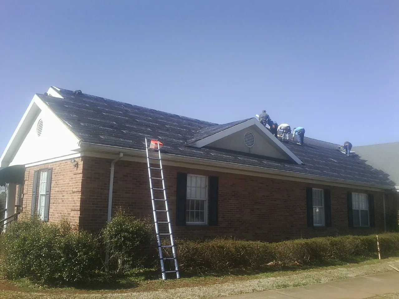 Crew tearing shingles off a brick ranch roof with a ladder set — Emergency Roof Repair work by our crew in Ronco