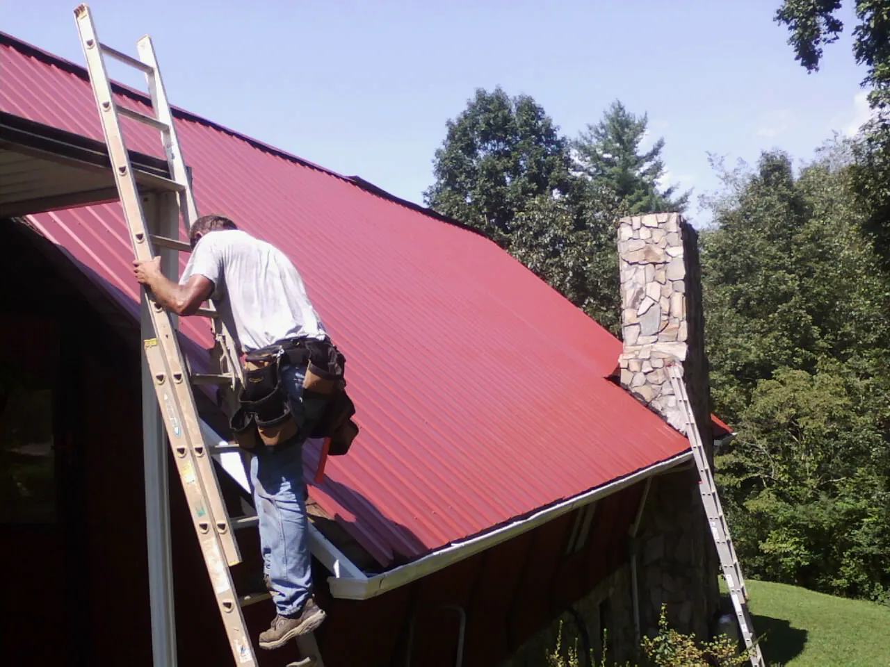 Roofer on a ladder beside a red metal roof and stone chimney — Soffit & Fascia Repair by Roofing Brothers in Ronco